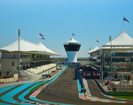 На изображении гоночная трасса с трибунами и контрольной башней. По бокам находятся оборудованные зоны с белыми навесами и флагами. Надписи на рекламных баннерах и голубое небо завершают картину