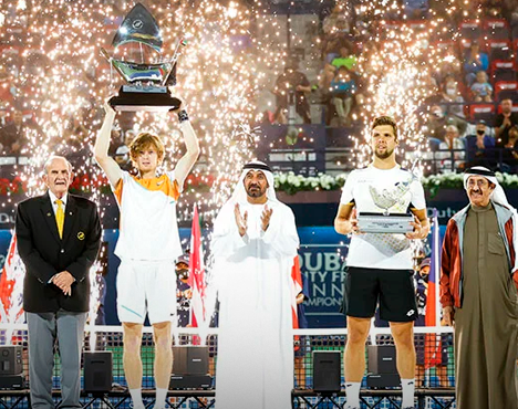 Победитель и финалист теннисного турнира на подиуме с трофеями. На заднем плане яркие спецэффекты с фейерверками. Рядом стоят официальные лица в традиционной и деловой одежде